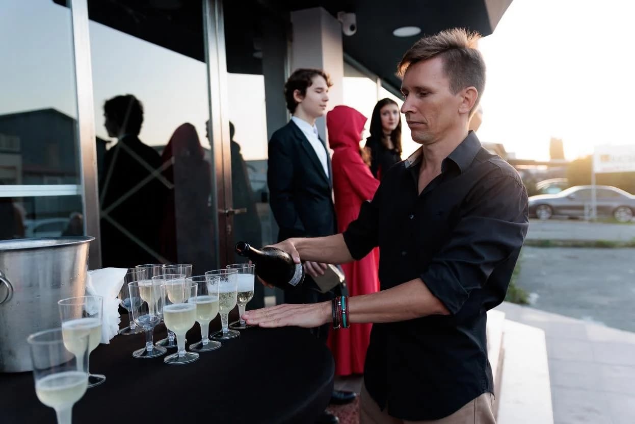 Andrew Sakhno pouring sparkling wine for guests at the entrance, actors in red behind him